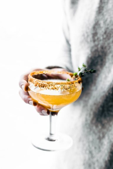 Cocktails/Mocktails Ein Glas mit orangefarbenem Cocktail, dekoriert mit einer Limettenscheibe und einem Zweig Thymian.